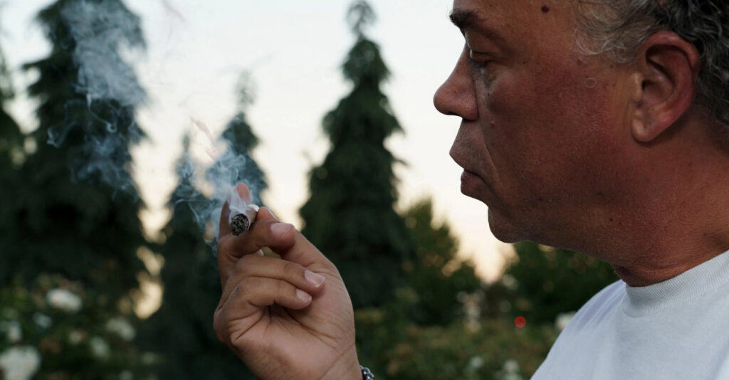 Joints, Blunts, and Spliffs: Understanding the Key Differences Profile of a man exhaling smoke from a hand-rolled blunt while standing outdoors with evergreen trees in the background.