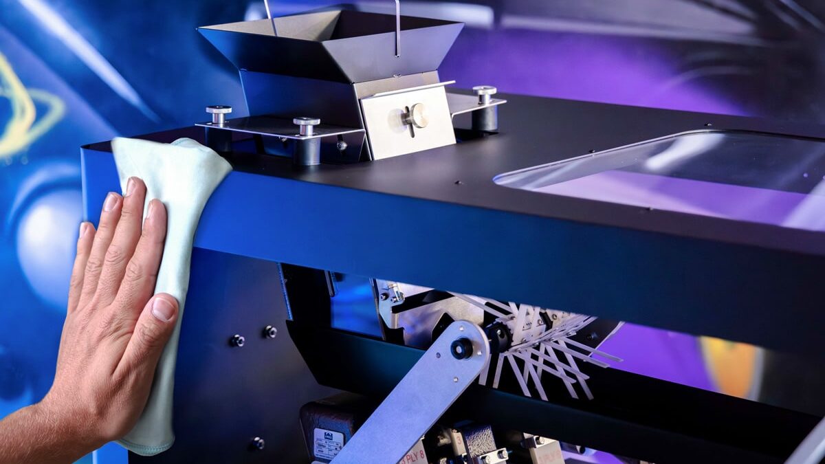 The 5 Biggest Pre-Roll Production Bottlenecks (And Why They're Probably Not Your Machine) A hand wiping down a blue automated production machine with a cloth under purple and blue industrial lighting.